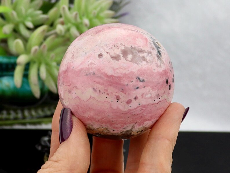May include: A polished, spherical gemstone with a swirling pattern of pink and white hues. The stone has small black inclusions and a smooth, rounded shape. The gemstone is held in a hand with a dark purple nail polish.