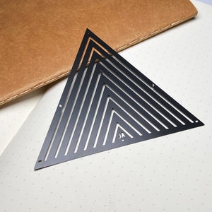 May include: A black metal triangle stencil featuring a series of nested triangles, forming a striped design. The stencil rests on dotted paper, with a brown leather notebook in the background. The initials "JA" are visible on the stencil.