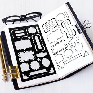 Peut inclure: Carnet ouvert avec des feuilles d'autocollants noirs et blancs présentant diverses formes et motifs d'étiquettes. Un stylo doré et une pince à reliure sont visibles. Des lunettes noires reposent sur le dessus du carnet. Le texte "JAYBEN'S APPLE" est imprimé.