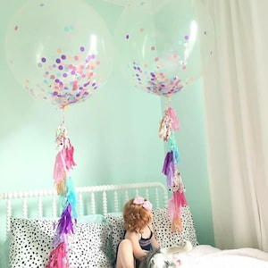 Toddler sitting on a bed with two 3ft clear balloon stuffed with tissue confetti and tassel string.