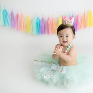 A little baby sits in a mint tutu clapping and smiling in front of a pastel themed garland strung across the wall behind. The colors of the garland are pink, peach, light yellow, light blue, mint, and lilac.