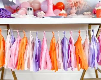 May include: A festive Halloween party scene with a pink cake stand holding a purple cake decorated with bats. A garland of colorful paper tassels in orange, pink, and purple hangs in the foreground. Balloons in pink, orange, and purple decorate the background.