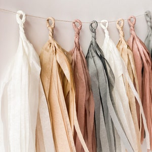 A close up side view of soft boho themed tissue tassel garland strung on a white wall shows the colors cream, tan, mauve, and grey.