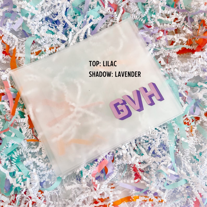 May include: A clear plastic bag with the text "TOP: LILAC SHADOW: LAVENDER" and the letters "GVH" in a 3D purple font. The bag is sitting on a bed of white, orange, pink, and blue shredded paper.