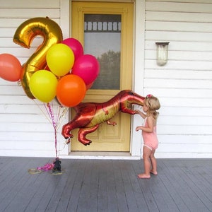 A little girl on the front porch of a house with a bouquet of balloons in yellow, coral, orange, wildberry, a number 2 in gold, and dinosaur balloon.