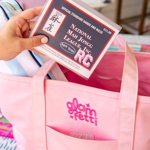 May include: A pink tote bag with the embroidered text "glam fetti" and a front pocket. A clear plastic sleeve with the text "NATIONAL MAH JONGG LEAGUE, INC." and "2025" is held above the bag. The bag contains a striped blue and white pouch.