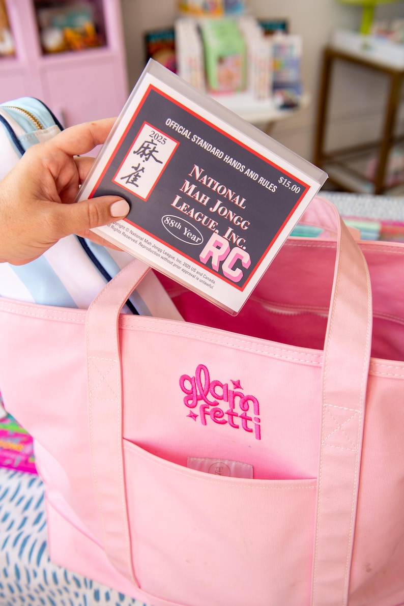 May include: A pink tote bag with the embroidered text "glam fetti" and a front pocket. A clear plastic sleeve with the text "NATIONAL MAH JONGG LEAGUE, INC." and "2025" is held above the bag. The bag contains a striped blue and white pouch.
