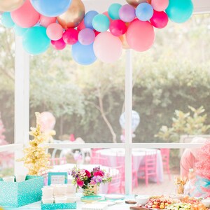 A vibrant balloon garland hangs draped from a ceiling above a table holding a few flower bouquets and a charcuterie board. The colors in the balloon garland are wildberry, periwinkle, Caribbean blue, rose, spring lilac, and chrome gold.