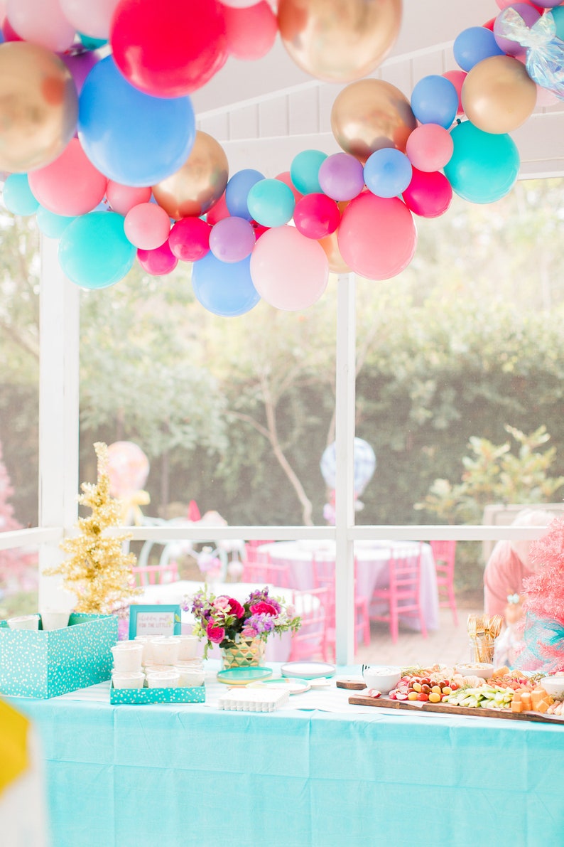 A vibrant balloon garland hangs draped from a ceiling above a table holding a few flower bouquets and a charcuterie board. The colors in the balloon garland are wildberry, periwinkle, Caribbean blue, rose, spring lilac, and chrome gold.