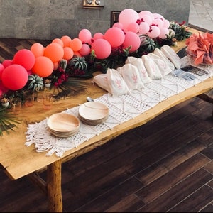 A balloon garland sits atop a bed of florals running along the middle of a wooden table.  The balloon garland is made up of wildberry, coral, rose, and pink balloons.