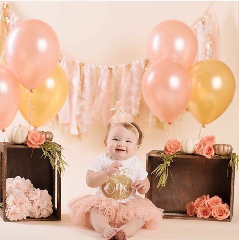 A little girl sitting on the ground with rose gold and gold balloons floating next to her.