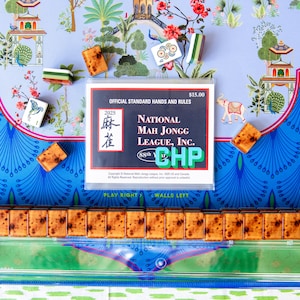 May include: A colorful image of a Mahjong set with tiles, a clear acrylic rack, and a rule book. The tiles are brown and rectangular. The rule book is white with black and red text. The background has a blue and green floral pattern.