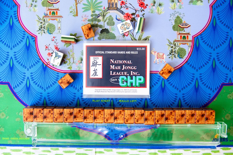 May include: A colorful image of a Mahjong set with tiles, a clear acrylic rack, and a rule book. The tiles are brown and rectangular. The rule book is white with black and red text. The background has a blue and green floral pattern.