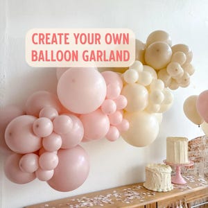 May include: A balloon garland featuring various sizes of matte balloons in shades of soft pink and cream. A rectangular sign with a light background and pink text reads CREATE YOUR OWN BALLOON GARLAND. Two frosted cakes sit on a wooden surface below.