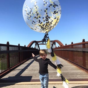 Little boy holding a jumbo 3ft clear balloon filled with tissue confetti in the colors white, black, and yellow with matching balloon string tassels.