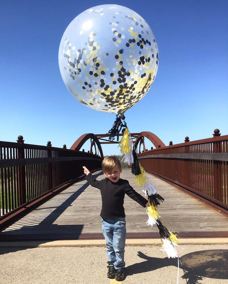 Little boy holding a jumbo 3ft clear balloon filled with tissue confetti in the colors white, black, and yellow with matching balloon string tassels.