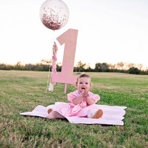 Toddler girl sitting on a blanket with a giant 3ft clear balloon stuffed with tissue confetti in the background next to a giant number one sign.