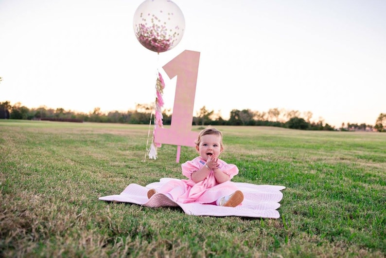 Toddler girl sitting on a blanket with a giant 3ft clear balloon stuffed with tissue confetti in the background next to a giant number one sign.