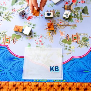 May include: A close-up of a Mahjong game in progress. The game features brown tiles with a speckled design, and white and green tiles. The game is set on a floral patterned surface with the initials "KB" visible.