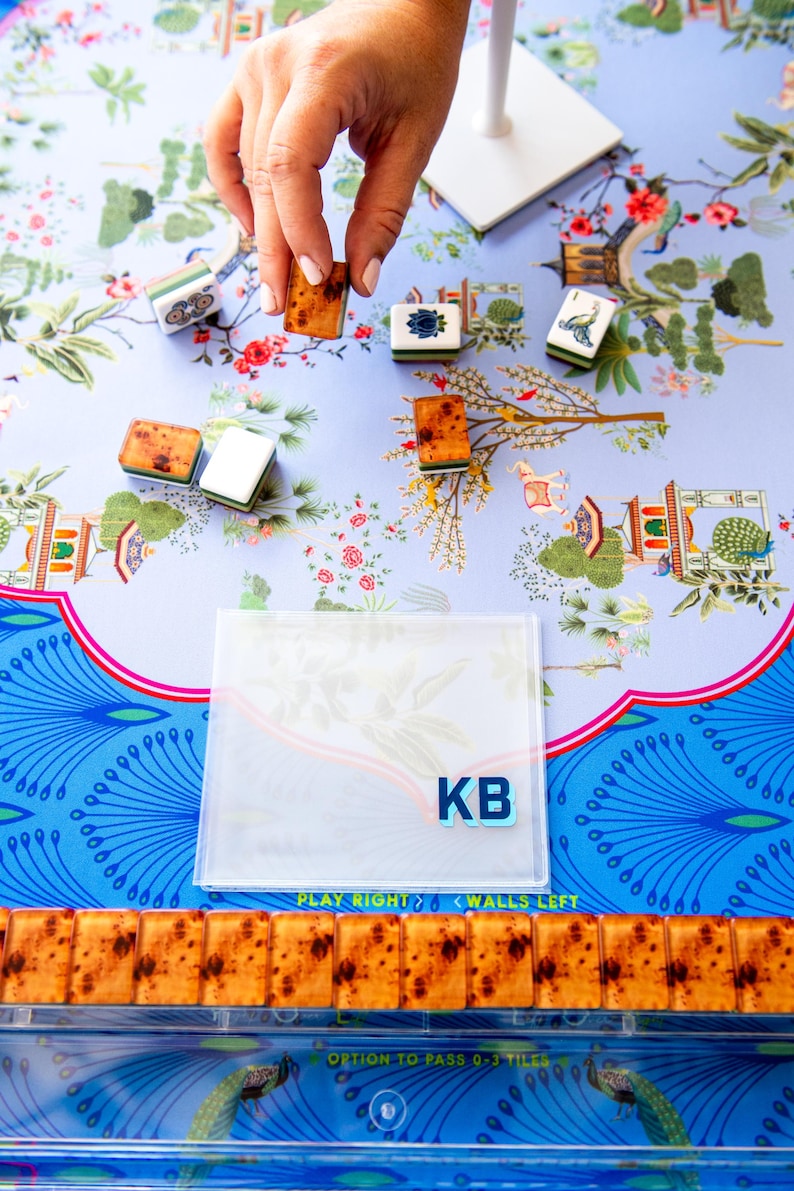 May include: A close-up of a Mahjong game in progress. The game features brown tiles with a speckled design, and white and green tiles. The game is set on a floral patterned surface with the initials "KB" visible.
