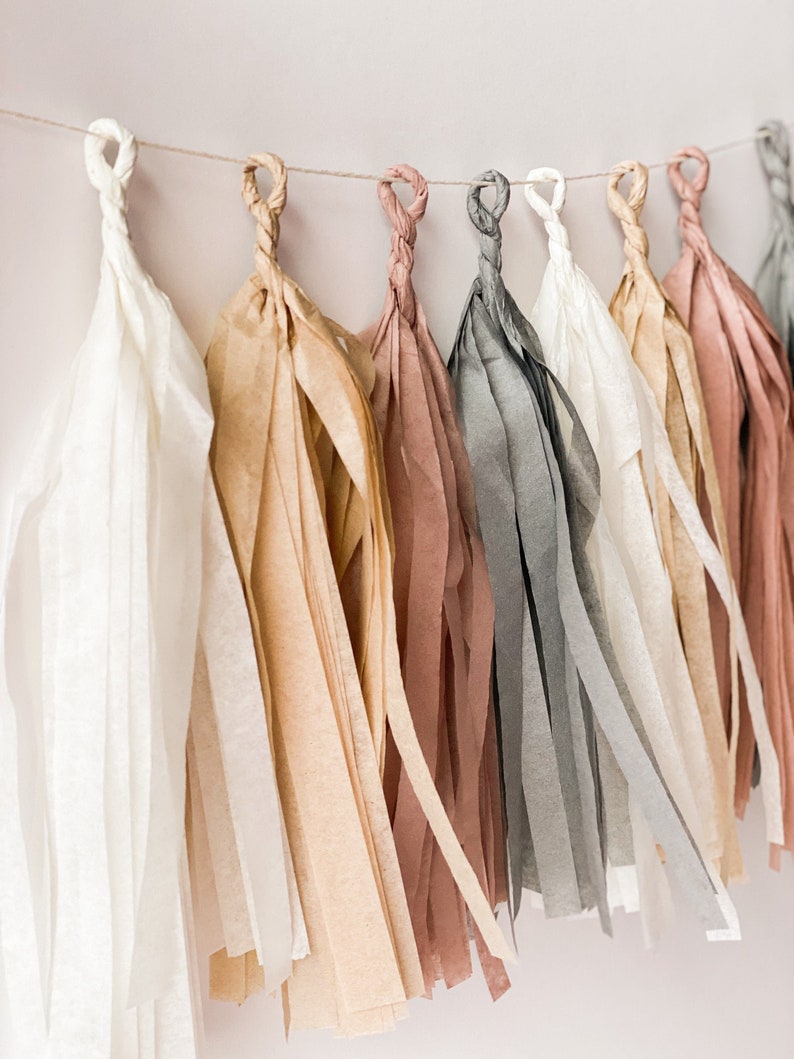 A close up side view of soft boho themed tissue tassel garland strung on a white wall shows the colors cream, tan, mauve, and grey.