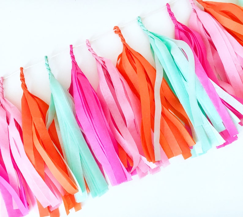 A arial shot of a hot pink, pink, orange, and mint tissue tassel garland is taken of tassels strung on twine and laying on a white table. The tassels are approximately nine inches long.