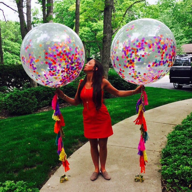 Woman holding two 3ft jumbo clear balloons stuffed with tissue confetti in the colors purple, pink, red, yellow, and orange and matching tassel tail.