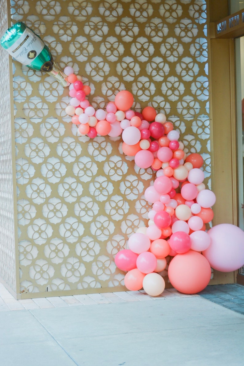 A tall wall with a champagne bottle balloon at the top left corner and pink, coral, and dark pink balloons in different sizes cascading out of the champagne bottle balloon.