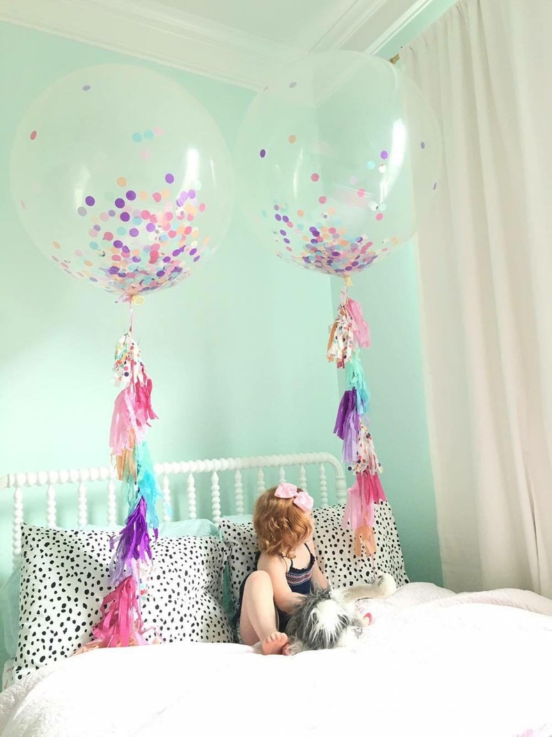 Toddler sitting on a bed with two 3ft clear balloon stuffed with tissue confetti and tassel string.