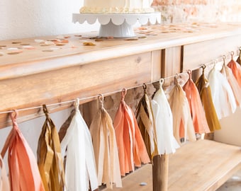 Puede incluir: Una mesa de madera con una tarta blanca sobre un soporte para tartas festoneado. Una guirnalda de borlas de papel en tonos melocotón, dorado y blanco cuelga delante. Confeti salpica la superficie de la mesa.
