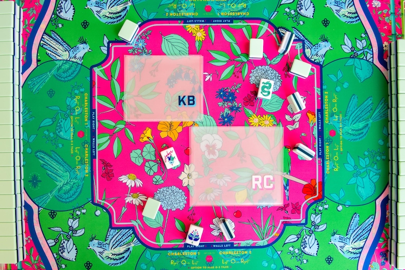 May include: A colorful Mahjong game set with a vibrant floral and bird design. The game board features a pink center with the initials "KB" and "RC" on clear covers. Green and white tiles are visible, along with floral and bird motifs.