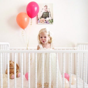 A little girl in a white crib holding a wildberry and orange balloon inflated with helium.