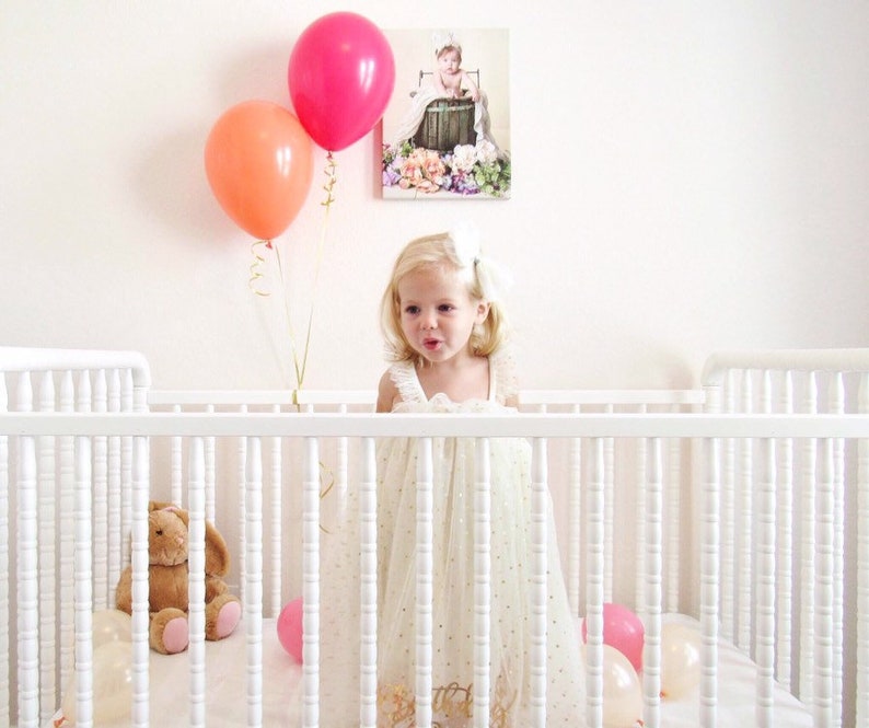 A little girl in a white crib holding a wildberry and orange balloon inflated with helium.