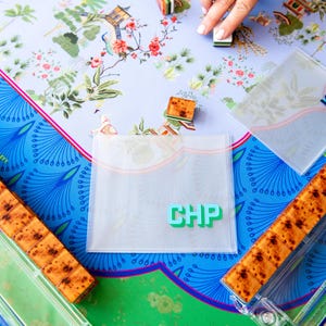 May include: A colorful game board with a floral design, featuring a light blue background and a vibrant green and blue border. The game includes wooden tiles, clear plastic holders, and a clear plastic pouch with the letters "CHP".