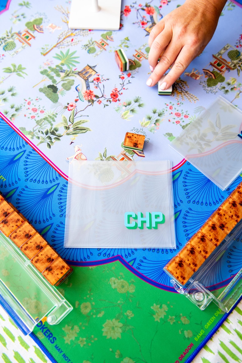 May include: A colorful game board with a floral design, featuring a light blue background and a vibrant green and blue border. The game includes wooden tiles, clear plastic holders, and a clear plastic pouch with the letters "CHP".