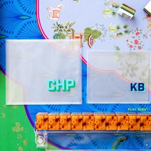 May include: Overhead shot of a colorful game setup. Two clear square cases with the letters "CHP" and "KB" are visible. The game pieces are arranged on a patterned surface with floral and peacock designs. The game appears to be Mahjong.