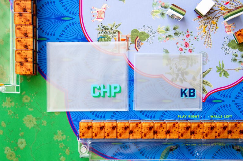 May include: Overhead shot of a colorful game setup. Two clear square cases with the letters "CHP" and "KB" are visible. The game pieces are arranged on a patterned surface with floral and peacock designs. The game appears to be Mahjong.