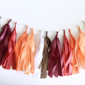 An arial view of a brown, maroon, red, orange, peach, and white tissue tassel garland sitting on a white table.
