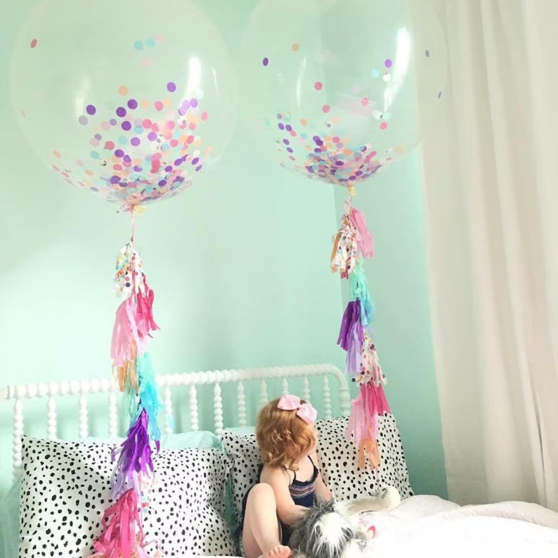 Puede incluir: Dos globos grandes transparentes llenos de confeti de colores y atados con largas serpentinas de borlas arco&iacute;ris. Los globos est&aacute;n en un dormitorio con una cama blanca y un ni&ntilde;o sentado en la cama.