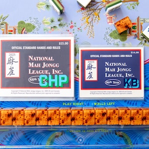 May include: A still-life image of Mahjong game components. Two rule books are visible, along with several tiles. The books are labeled "National Mah Jongg League, Inc." and are dated 2023 and 2025. The tiles are brown and white.