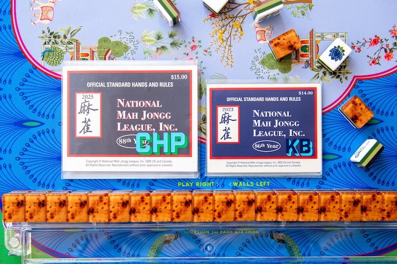 May include: A still-life image of Mahjong game components. Two rule books are visible, along with several tiles. The books are labeled "National Mah Jongg League, Inc." and are dated 2023 and 2025. The tiles are brown and white.