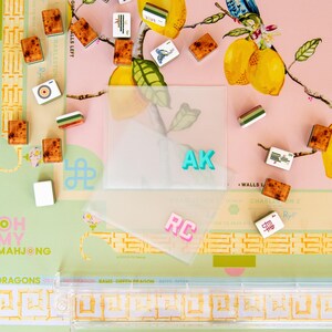 May include: A Mahjong game set with tiles scattered on a pink and green floral patterned surface. The set includes tiles with various symbols and colors, along with clear plastic card holders with the initials "AK" and "RC".