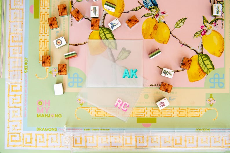 May include: A Mahjong game set with tiles scattered on a pink and green floral patterned surface. The set includes tiles with various symbols and colors, along with clear plastic card holders with the initials "AK" and "RC".