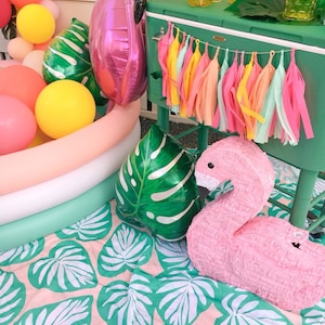 A flamingo pool party scene shows a tissue tassel garland strung across the front of a tropical green cooler. The colors in the garland are coral, hot pink, yellow, aqua, and peach.