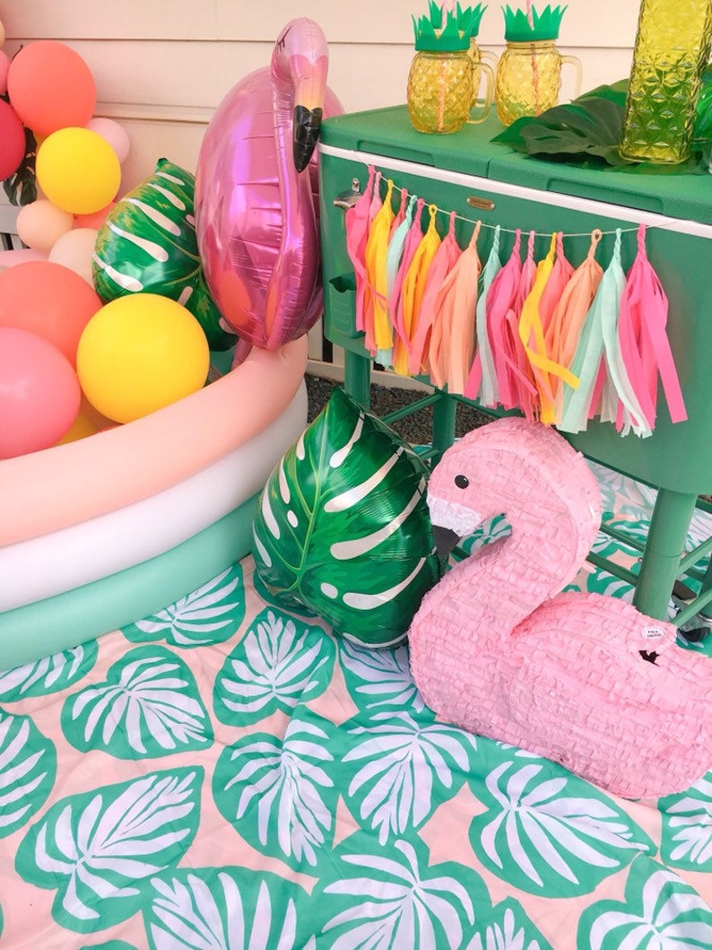 A flamingo pool party scene shows a tissue tassel garland strung across the front of a tropical green cooler. The colors in the garland are coral, hot pink, yellow, aqua, and peach.