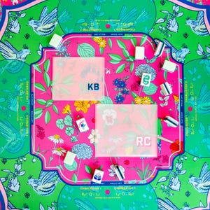 May include: A colorful Mahjong set with a floral and bird design. The set includes tiles, racks, and a playing surface with pink, green, and blue accents. The playing surface has the initials "KB" and "RC".