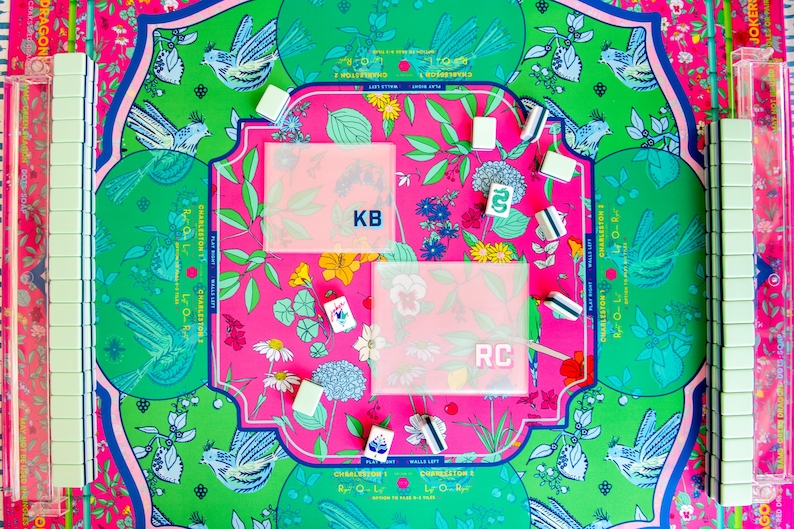 May include: A colorful Mahjong set with a floral and bird design. The set includes tiles, racks, and a playing surface with pink, green, and blue accents. The playing surface has the initials "KB" and "RC".