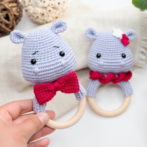 May include: Two handmade crochet hippo baby rattles. The hippos are light grey with black eyes and a red bow tie or scarf. The rattles have wooden rings. One hippo has a white flower on its head.