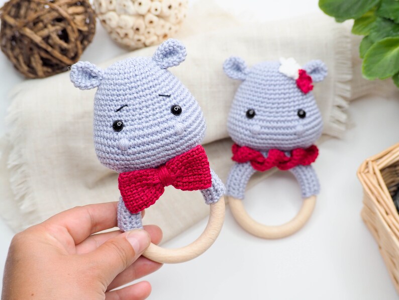 May include: Two handmade crochet hippo baby rattles. The hippos are light grey with black eyes and a red bow tie or scarf. The rattles have wooden rings. One hippo has a white flower on its head.