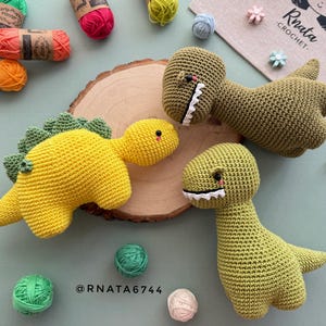 May include: Crocheted dinosaur toys in yellow and green, with white teeth and red accents. The toys are displayed with colorful yarn balls and a wooden slice. The image includes the text "Rnata Crochet" and "@RNATA6744".